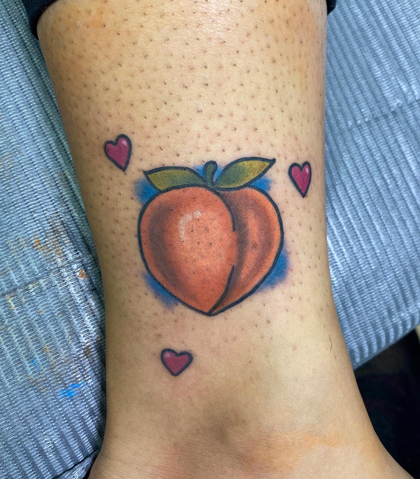 Ankle tattoo of a peach with green leaves and three small pink hearts on skin